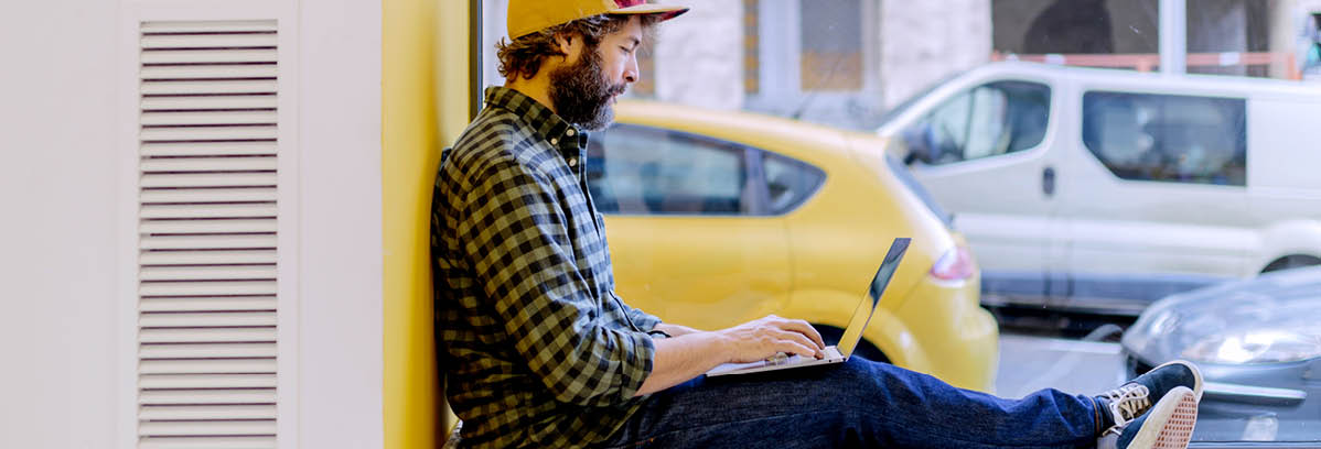 Bearded man in cap and checkered shirt typing on laptop sitting on window sill against yellow wall