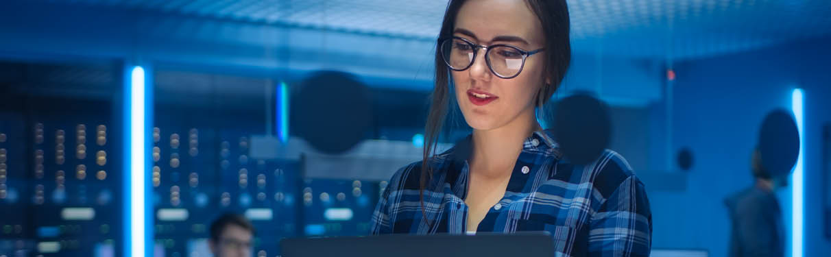 Portrait of a Smart Seductive Young Woman Wearing Glasses Holds Laptop. In the Background Technical Department Office with Specialists Working and Functional Data Server Racks