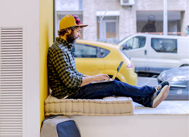 Bearded man in cap and checkered shirt typing on laptop sitting on window sill against yellow wall