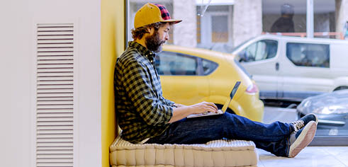 Bearded man in cap and checkered shirt typing on laptop sitting on window sill against yellow wall