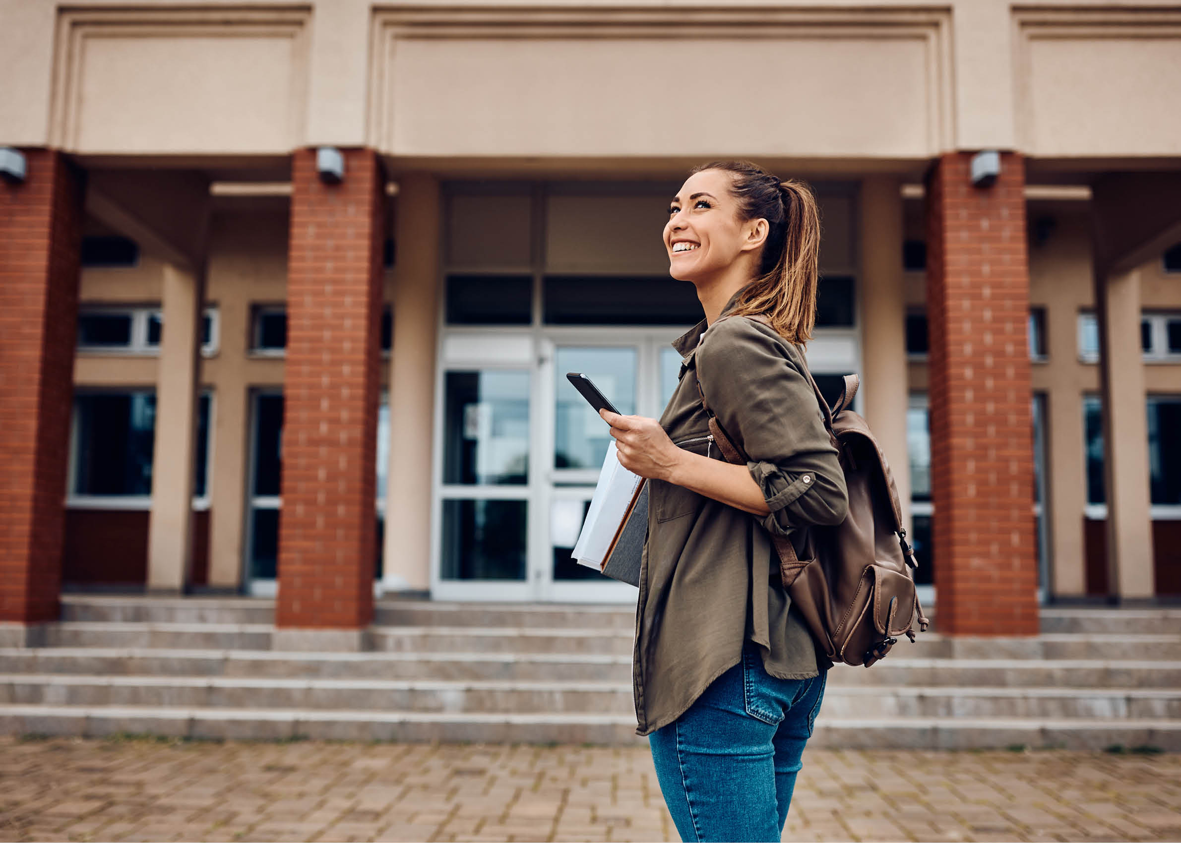 Happy female student texting on smart phone while going to lecture at the university. Copy space. 