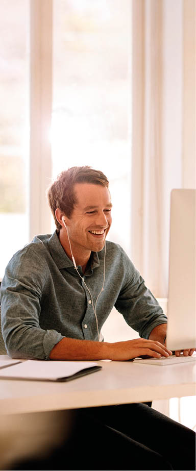 Smiling businessman working on computer. Man sitting at his work table working on computer at home listening to audio using earphones.
