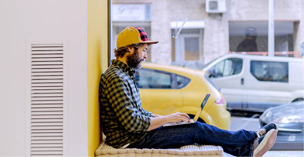 Bearded man in cap and checkered shirt typing on laptop sitting on window sill against yellow wall